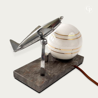 English: Vintage Art Deco desk lamp with a polished chrome Caudron Simoun airplane in flight position mounted on a marble base, next to a glowing white opaline sphere with gold stripes. Homage to Antoine de Saint-Exupéry, circa 1937.
French: Lampe de bureau Art Déco vintage avec un avion Caudron Simoun chromé en position de vol, monté sur une base en marbre à côté d’un globe opaline blanc à rayures dorées. Hommage à Antoine de Saint-Exupéry, vers 1937.