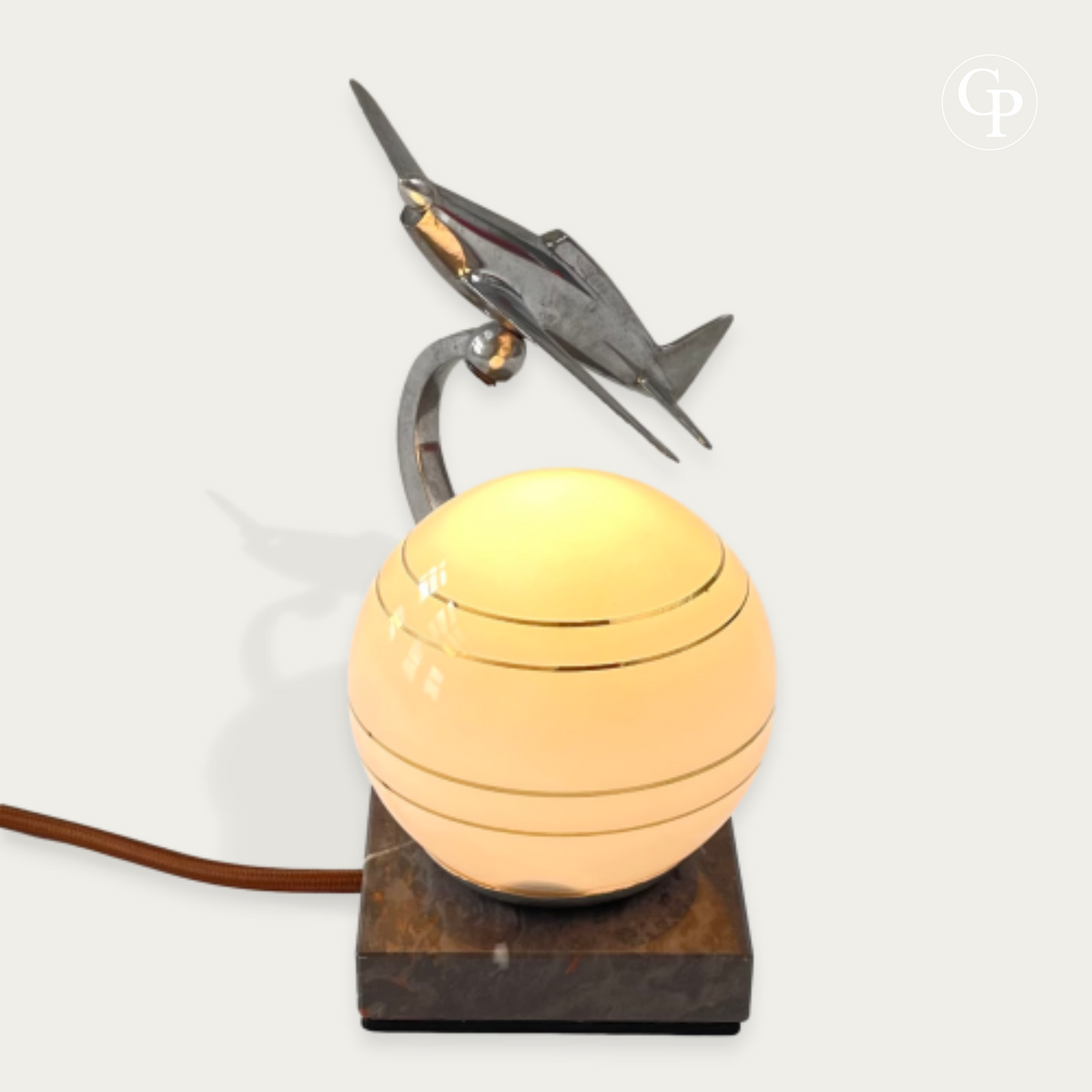 Beautifully lit vintage Art Deco table lamp with a glowing warm opaline sphere and a chrome Caudron Simoun airplane in dynamic ascending flight position on a marble base. Homage to Antoine de Saint-Exupéry, circa 1937.