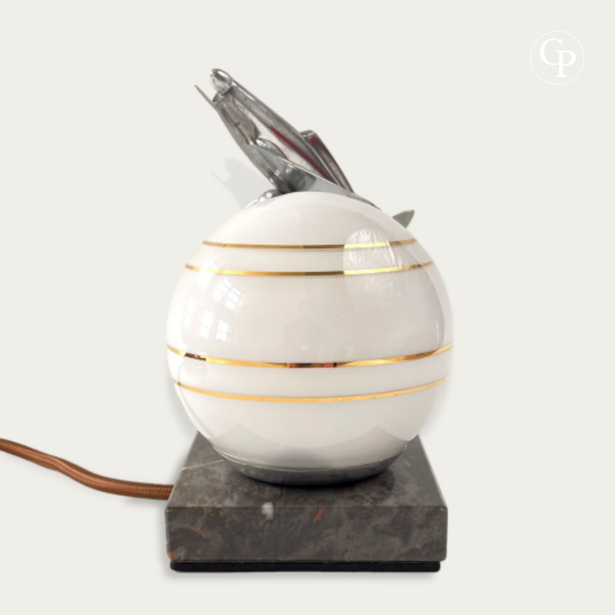 English: Vintage Art Deco table lamp featuring a chrome Caudron Simoun airplane on a glowing white opaline sphere with gold stripes, mounted on a marble base. Homage to Antoine de Saint-Exupéry, circa 1937. | Français : Lampe de table Art Déco vintage avec un avion Caudron Simoun chromé posé sur un globe opaline blanc à fines rayures dorées, sur base en marbre. Hommage à Antoine de Saint-Exupéry, vers 1937.