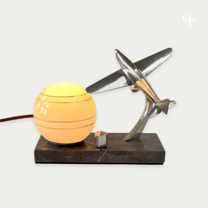 English: Vintage Art Deco table lamp with warm glowing opaline sphere and a polished chrome Caudron Simoun airplane in dynamic flight position on a marble base. Homage to Antoine de Saint-Exupéry, circa 1937. | French: Lampe de table Art Déco vintage avec globe opaline lumineux à lumière chaude et avion Caudron Simoun chromé en position de vol dynamique sur base en marbre. Hommage à Antoine de Saint-Exupéry, vers 1937.