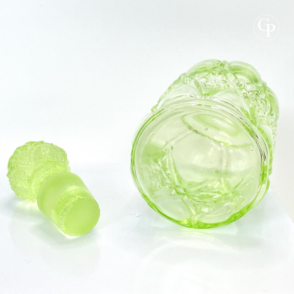 Side view of the base of the rare Val Saint Lambert Valambert n°12 Ouraline uranium glass perfume bottle with its original pineapple stopper placed beside it, showcasing the thick glass and craftsmanship, Art Deco circa 1935. | Vue de côté sur le fond du rare flacon Val Saint Lambert Valambert n°12 en verre à l'uranium Ouraline avec son bouchon d'origine en forme de pomme de pin posé à côté, mettant en valeur l'épaisseur du verre et la qualité d'exécution, Art Déco vers 1935.