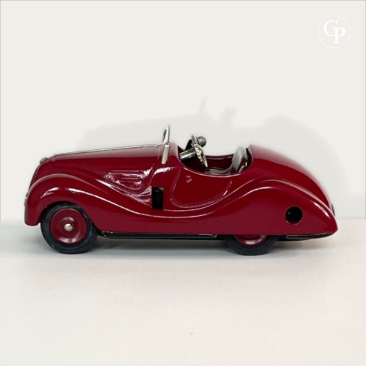 Full side profile view of the deep red Schuco Examico 4001 vintage tinplate roadster, highlighting the sleek aerodynamic design, windshield, and rubber tires on a light background. | Vue de profil complet de la voiture automate Schuco Examico 4001 couleur bordeaux, mettant en valeur sa silhouette aérodynamique, son pare-brise et ses pneus en caoutchouc sur fond clair.